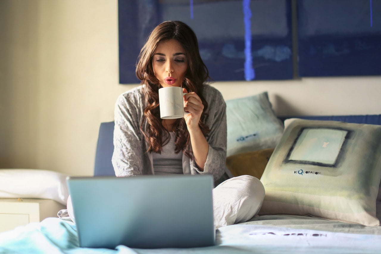 Image of a woman sitting on her bed drinking a cup of coffee while on lapto